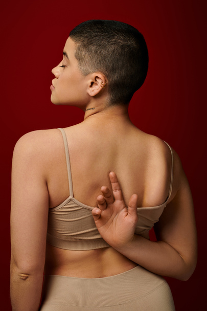 body positive, self-esteem, tattooed young woman with short hair and tattoo posing with hand behind back on burgundy background, dark red, curvy fashion, comfortable in skin, female underwear  - Foto, Imagen