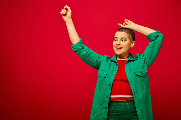 fashion and style, happy and short haired woman in green outfit posing with raised hands on red background, generation z, youth culture, modern backdrop, individuality, personal style  - Photo, Image
