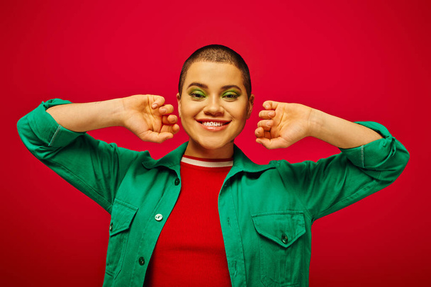 fashion statement, cheerful and short haired woman in green outfit posing on red background, generation z, youth culture, modern backdrop, individuality, personal style, looking at camera - Photo, Image