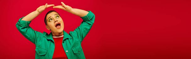 fashion and style, amazed and short haired woman in green outfit posing with raised hands on red background, looking up generation z, youth culture, vibrant backdrop, personal style, banner  - Фото, зображення