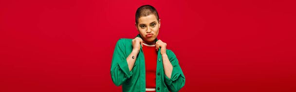 fashion choices, emotional and tattooed, short haired woman in green outfit pouting lips on red background, looking at camera, generation z, youth culture, vibrant backdrop, style, banner  - Photo, Image