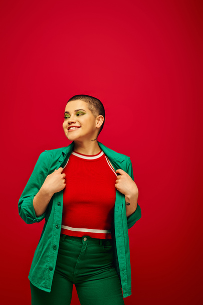 fashion trend, happy and tattooed, short haired woman in green outfit smiling on red background, looking away, generation z, youth culture, vibrant backdrop, individuality, personal style - Photo, Image