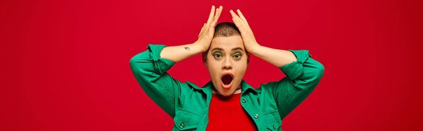 fashion trend, shocked and tattooed, short haired woman in green outfit touching head on red background, looking away, generation z, youth, vibrant backdrop, individuality, personal style, banner  - 写真・画像