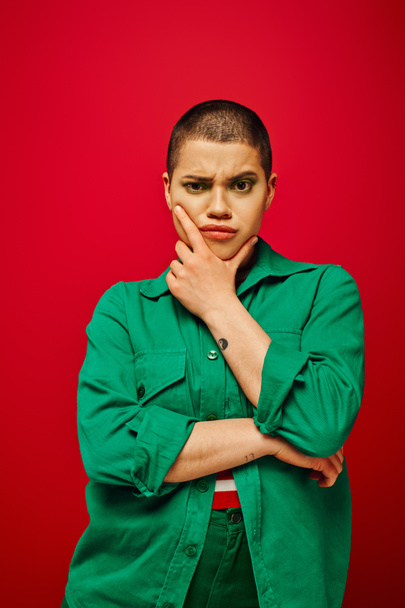 fashion choices, displeased and tattooed, short haired woman in green outfit touching chin on red background, looking at camera, generation z, youth, vibrant backdrop, individuality, personal style  - Photo, Image