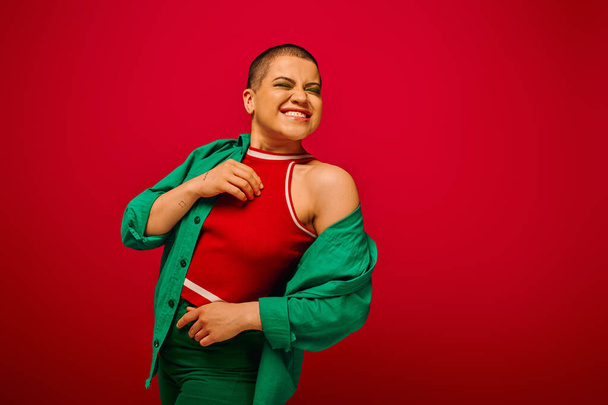 fashion statement, bold makeup, pleased and tattooed, short haired woman in green outfit smiling on red background, generation z, youth, vibrant backdrop, individuality, personal style  - Photo, Image