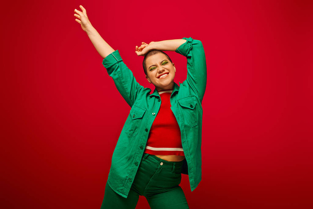 stylish outfit, bold makeup, cheerful and tattooed, short haired woman in green outfit posing on red background, generation z, youth culture, vibrant backdrop, personal style  - Valokuva, kuva