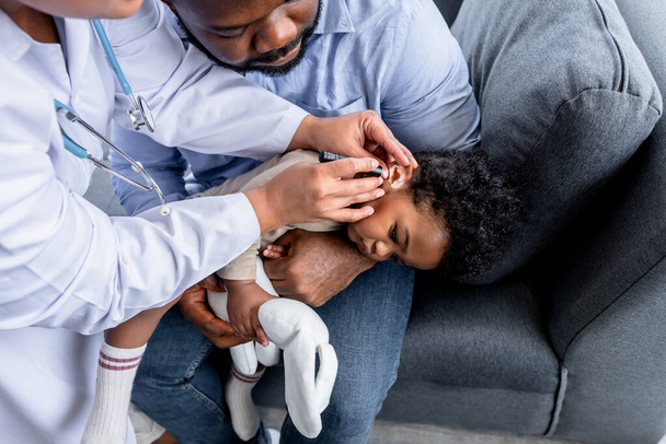 Жіночий лікар робить попередній екзамен на вухо 10-місячного хлопчика нігерійського малюка, чий батько тримає його. до медичного догляду немовлятко й слухове зараження. - Фото, зображення