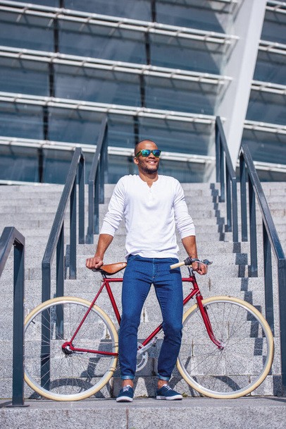 Full length portrait of handsome young businessman in casual wear and sun glasses smiling and leaning on bike while standing on stairs outdoors - Photo, Image