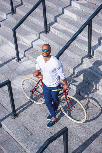 Blick aus der Vogelperspektive auf einen gut aussehenden jungen Geschäftsmann in Freizeitkleidung und Sonnenbrille, der sich auf sein Fahrrad stützt, während er im Freien auf einer Treppe steht - Foto, Bild