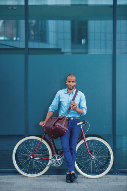 Retrato de larga duración de un joven hombre de negocios guapo con ropa casual usando un teléfono inteligente y mirando a la cámara mientras se apoya en su bicicleta, de pie al aire libre - Foto, Imagen