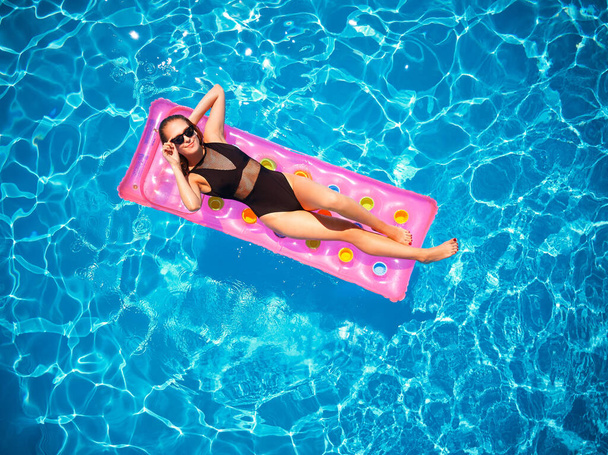 Fit pretty girl in bikini chilling on inflatable pink mattress in swimming pool. Slim hot woman in swimwear tanning. Female relaxing on a float in blue water at luxury resort. Aerial, view from above. - Fotografie, Obrázek