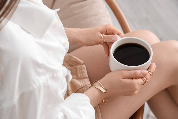 Mujer joven sentada en sillón y sosteniendo una taza de delicioso café - Foto, Imagen