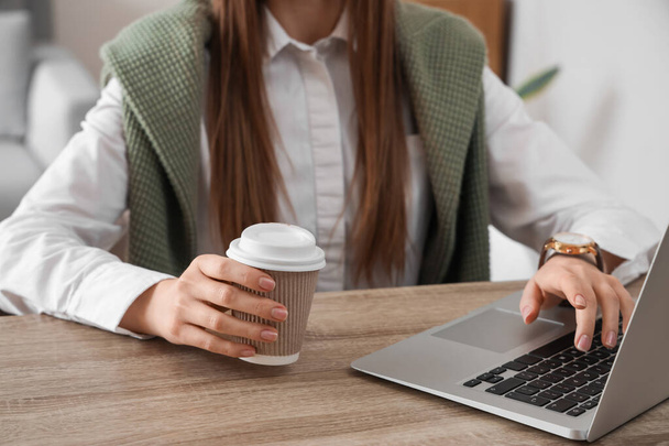 Жінка використовує сучасний ноутбук і тримає чашку кави за столом
 - Фото, зображення