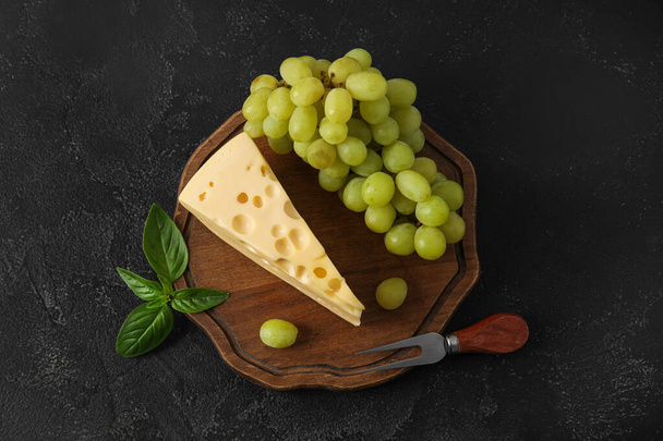 Houten plank met lekkere Zwitserse kaas en druiven op zwarte ondergrond - Foto, afbeelding