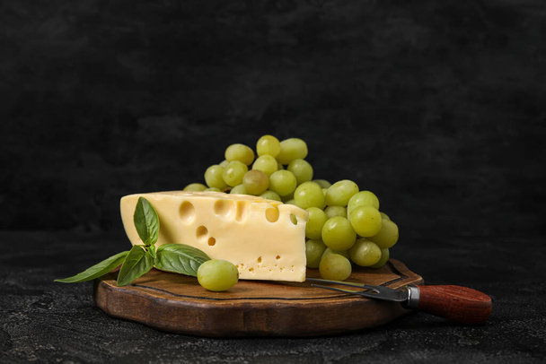 Wooden board with tasty Swiss cheese and grapes on black background - Valokuva, kuva