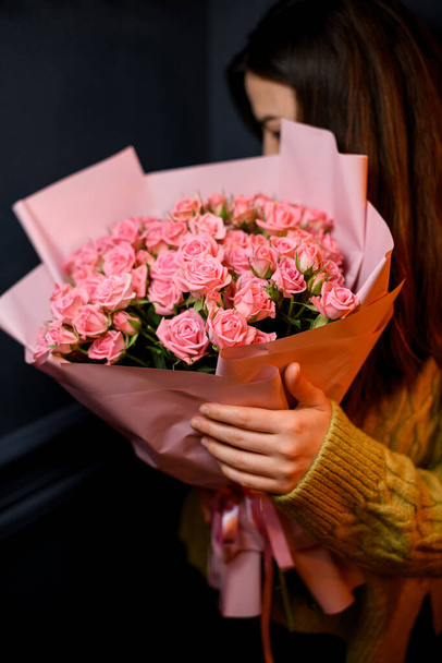 Young darkhair woman florist holding in hands large bouquet of fresh pink roses wrapped in pink paper and decorated with satin ribbon - Foto, Bild