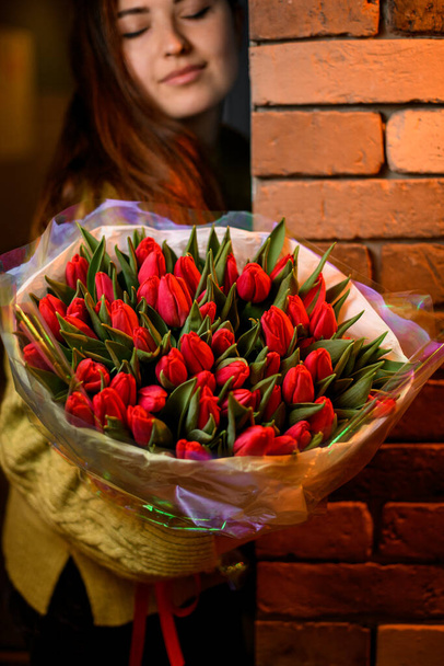 Woman florist holds bouquet of fresh red tulips, close her eyes enjoying beautiful flovers. Large, beautiful, fragrant bouquet of flowers - Photo, Image