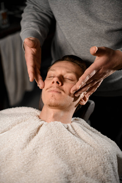 Barber in barbershop making soft massage of clients face to prepare it for shaving process. Gentle shave, beard care for men, male beauty and care concept - Photo, image