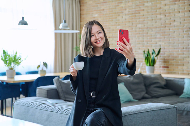 Young woman having video call chat conference sitting on sofa at home, using smartphone. Female recording video blog vlog making online broadcast talking with friends, leisure study work lifestyle - Фото, зображення