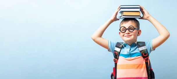 Divertido niño sonriente escolar en gafas sosteniendo libros en su cabeza sobre fondo azul - Foto, Imagen