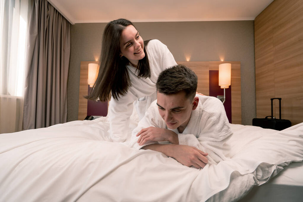 retrato joven pareja en luna de miel en hotel habitación acostado cama en blanco batas coqueteando riendo feliz amantes - Foto, Imagen