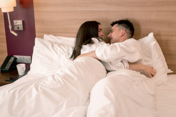 Balayında genç bir çiftin portresi otel odasında beyaz cüppeler üzerinde uzanıp mutlu aşıklar için seyahat kavramını öpüyor. - Fotoğraf, Görsel