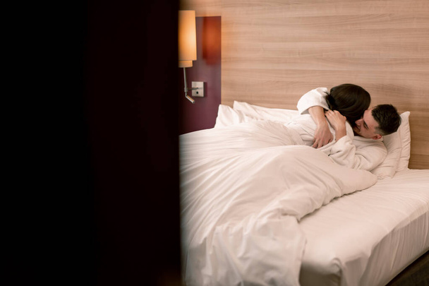 портрет молодої пари на медовий місяць в готельному номері лежить на ліжку білі халати поцілунки щасливі коханці концепція подорожей - Фото, зображення