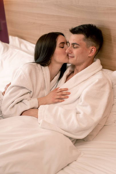 portrait young couple on honeymoon in hotel room lying on bed white robes kissing happy lovers travel concept - Foto, afbeelding