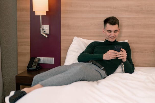 ένας νεαρός στο δωμάτιο ενός πολυτελούς ξενοδοχείου κρατάει ένα smartphone στα χέρια του και λύνει επαγγελματικά προβλήματα μετά το ταξίδι - Φωτογραφία, εικόνα
