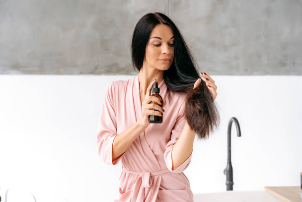 Hair spray. Female beauty routine. Hair care concept. Beautiful brunette caucasian woman applying essential oil spray on her long hair while sitting in a bathroom, smiling. Split ends repair treatment - Photo, image
