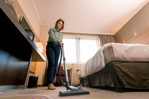 νεαρή κοπέλα οικιακή βοηθός στο δωμάτιο καθαρισμού κορίτσι του ξενοδοχείου σκούπισμα χαλί στο πολυτελές δωμάτιο έννοια καθαριότητα και φιλοξενία - Φωτογραφία, εικόνα