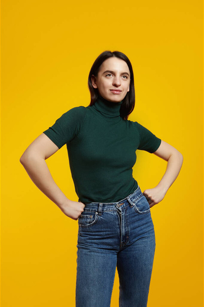 Vertical photo of young confident girl, wearing green t-shirt and jeans, holding fists on waist, shows her power. - Фото, изображение
