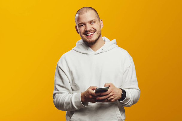 Cheerful unshaven man using mobile phone, wearing hoodie and smiling at camera, isolated over yellow background - Photo, image