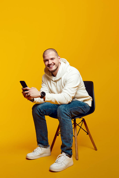 Good looking man using smartphone while sitting on chair isolated on yellow background. Excited casual guy chatting online, browsing social media - Foto, Imagen