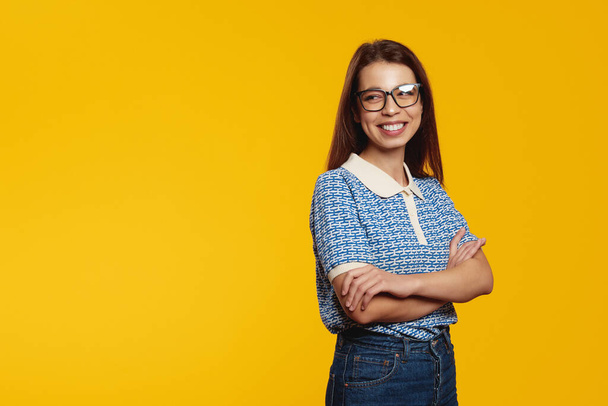 Chica joven feliz en camisa azul y anteojos sonriendo mientras mira el espacio libre vacío para el texto con brazos cruzados, aislado sobre fondo amarillo - Foto, imagen