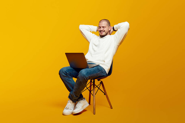 Guapo joven que se relaja en la silla y el uso de la computadora portátil, feliz hombre milenario inclinándose hacia atrás, mirando a la pantalla del ordenador portátil, aislado sobre el fondo amarillo - Foto, imagen