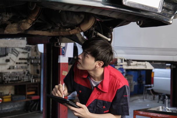 Professional young Asian male motor mechanic inspects undercarriage of electric car(EV) lifted by forklift jack for repair at garage, automotive maintenance service works industry occupation business. - Foto, Imagem