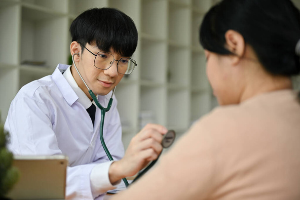 Un medico maschio asiatico millenario felice e professionale utilizza uno stetoscopio per esaminare il battito cardiaco di una paziente in ospedale. concetto di assistenza sanitaria - Foto, immagini