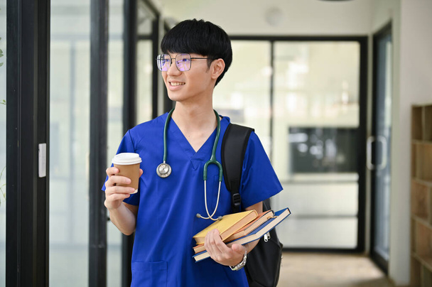 Onnellinen ja komea nuori aasialainen miespuolinen lääketieteen opiskelija tai lääkäri pensaikossa kävelee sairaalan käytävää pitkin kahvikuppi ja kirjat käsissään.. - Valokuva, kuva