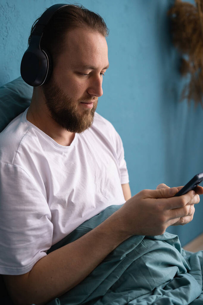 Усміхнений молодий бородатий чоловік лежить на ліжку вдома вранці, слухаючи музику за допомогою мобільного телефону та навушників. Копіювати простір
 - Фото, зображення