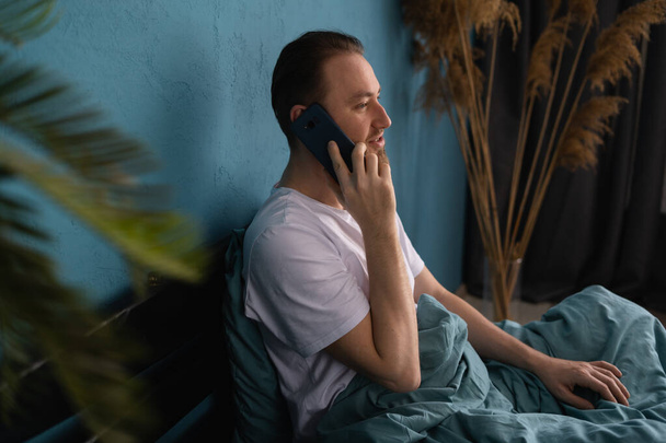 Komea mies puhuu matkapuhelussa sängyssä kotona aamulla. nauti viikonlopusta älypuhelimella teknologian kanssa. Kopioi tila - Valokuva, kuva