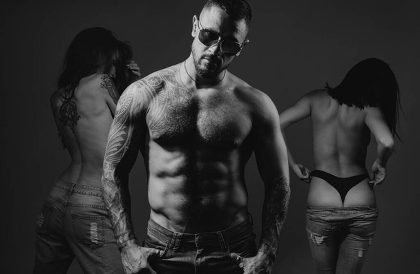 Sexy muscular macho man in sunglasses with two hot girls studio photo. Women with sexy fit ass standing behind handsome tattooed man with sexy torso - Fotografie, Obrázek