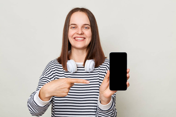 Attractive cheerful woman wearing striped shirt standing with headphones over neck isolated over gray background pointing at smartphone with empty display copy space for advertisement - Photo, Image