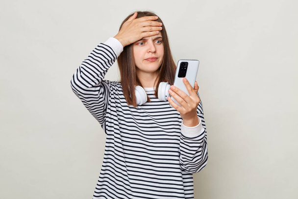 Shocked confused woman wearing striped shirt standing with headphones over neck isolated over gray background reading news in internet making facepalm gesture. - Photo, Image
