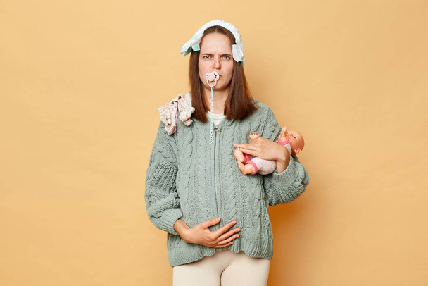 Sad upset pregnant woman wearing knitted warm sweater standing isolated over beige background holding doll and nipple looking at camera with displeased expression. - Photo, Image