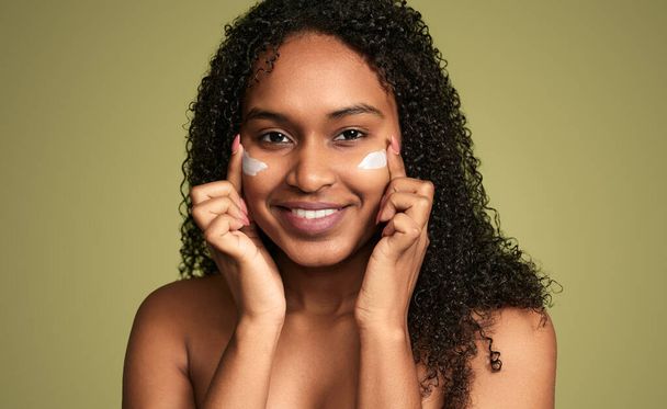 Content young African American female with flawless face skin smearing cream under eyes while looking at camera on green background - Фото, зображення