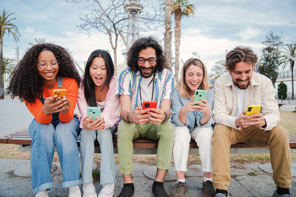 Grupa najlepszych przyjaciół korzystających i uśmiechnięty za pomocą aplikacji na telefon komórkowy siedzi na ławce udostępniania wiadomości. Szczęśliwi młodzi ludzie na czacie w mediach społecznościowych online z komórki zabawy razem - Zdjęcie, obraz