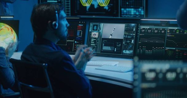 Διαφορετικοί χειριστές ελέγχου πτήσης σε ακουστικά παρακολουθούν διαστημική αποστολή σε υπολογιστές στο κέντρο ελέγχου αποστολής. Ομάδα παλαμάκια μετά την επιτυχή εκτόξευση διαστημικού πυραύλου εμφανίζεται σε μεγάλες ψηφιακές οθόνες. - Φωτογραφία, εικόνα