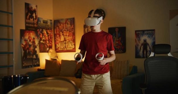 Niño en auriculares VR juega realidad aumentada videojuegos en línea utilizando controladores inalámbricos, pasa tiempo libre en el dormitorio por la noche. Concepto de dispositivos modernos, entretenimiento y estilo de vida. - Foto, imagen