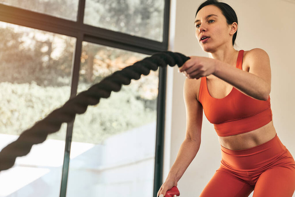 Low angle of fit female athlete in tight sportswear doing exercise with battle ropes during intense workout in modern fitness center - Foto, immagini